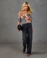 Woman wearing abstract print V-neck blouse with dark wide-leg jeans, black strappy heels, and woven clutch bag