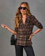 Woman wearing a brown plaid double-breasted blazer with black sunglasses, gold bangles, black quilted bag and satin pants
