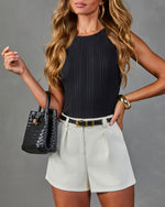 Woman wearing black ribbed sleeveless top, cream pleated shorts, black woven leather bucket bag, gold cuff bracelet and hoop earrings