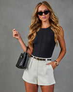 Woman wearing black ribbed tank top, cream pleated shorts, black belt, cat-eye sunglasses, holding woven leather bucket bag