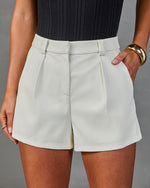 White high-waist pleated tailored shorts paired with dark ribbed bodysuit and gold cuff bracelet