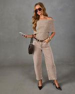 A woman models a beige off-the-shoulder knit top and matching beige wide-leg jeans with a brown belt, styled with black pointed-toe heels, sunglasses, and a small dark brown handbag against a gray studio backdrop.