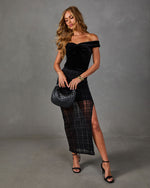 Woman in black velvet off-shoulder top and sheer plaid maxi skirt with high slit, holding woven handbag and black heeled sandals