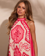 Woman wearing pink and red pleated halter maxi dress with medallion print, gold hoop earrings, wavy brown hair
