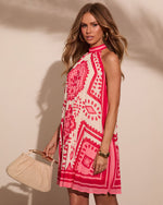 Woman wearing pink geometric print halter mini dress with pleated skirt, holding beige woven bag, gold jewelry, summer fashion