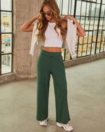 Woman wearing forest green wide-leg pants, white crop top, layered gold necklaces, aviator sunglasses, and chunky sneakers.
