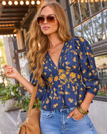 A woman wearing a navy blue floral blouse with yellow flowers, paired with light blue jeans, gold sunglasses, and a tan shoulder bag, standing in front of a storefront.