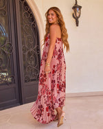Woman in pink floral pleated maxi dress and nude heels smiling over shoulder near ornate iron door