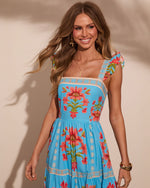 Woman wearing turquoise floral ruffle-strap sundress with pink lily print, smiling, gold jewelry, summer fashion