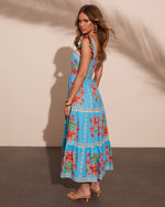 Woman modeling turquoise floral maxi dress with tiered skirt, smocked back, ruffle straps, and coral lily print, styled with gold heels