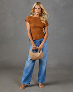 Woman in rust ribbed knit sweater top, wide-leg blue jeans, woven tan mini bag, and clear heeled sandals