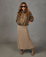 A woman modeling a brown patterned midi skirt with a small checkered print, styled with a brown jacket and sunglasses against a gray studio background.
