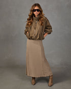 A woman modeling a brown patterned midi skirt with a small checkered print, styled with a brown jacket and sunglasses against a gray studio background.