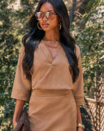 A woman wearing a tan two-piece set consisting of a V-neck wrap top with elbow-length sleeves and a matching pencil skirt, styled with gold hoop earrings, glasses, and layered necklaces against a blurred outdoor background.