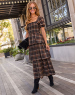 A woman wearing a brown and black plaid tiered midi dress with puff sleeves, styled with black ankle boots and holding a black studded clutch, posing on a paved walkway outside a building.