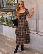 A woman models a brown and black plaid tiered midi dress with short puff sleeves and a square neckline, styled with black ankle boots, a black studded clutch, and sunglasses while leaning against a stone building.