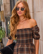 A woman wearing an off-the-shoulder brown plaid dress with puff sleeves, styled with gold-framed sunglasses and layered necklaces, standing against a stone wall.