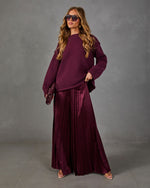 Woman in burgundy oversized knit sweater, satin pleated wide-leg pants, aviator sunglasses and clutch against gray backdrop