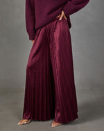 Burgundy satin pleated wide-leg pants styled with a plum knit sweater and clear-strap nude heels against a gray backdrop