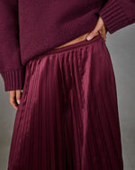 Burgundy satin pleated midi skirt styled with an oversized burgundy knit sweater, hand resting on hip, gray backdrop