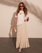 Woman wearing white crop tee, cream lace maxi skirt, white sandals, gold jewelry, and brown sunglasses holding beige mini bag
