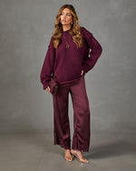 A woman modeling a matching burgundy knit sweater and wide-leg satin pants set with fringe hems, styled with a small matching clutch and clear high-heeled sandals against a gray studio backdrop.