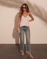 Woman wearing white lace cami top, low-rise wide-leg cuffed jeans, strappy heels, and sunglasses holding cream clutch
