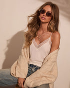 Woman in beige knit cardigan, white lace cami top, and jeans wearing gold sunglasses and layered necklaces
