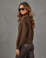 A woman wearing a brown knit mock-neck sweater and dark brown faux-leather pants, photographed from the back against a textured gray studio background.