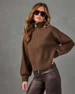 A woman wearing a brown ribbed knit zip-up sweater with a mock neck and long sleeves, paired with dark brown faux-leather pants, against a textured gray studio background.