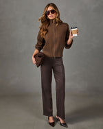 A woman models a brown knit zip-up sweater, dark brown wide-leg trousers, and black pointed-toe heels against a textured gray studio backdrop. She holds a brown clutch and a coffee cup.