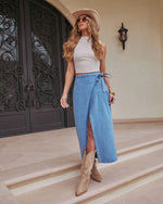 A woman wearing a white sleeveless top, tan cowboy boots, a beige hat, and the Maebelle Wrap Tie Denim Maxi Skirt stands before double black doors with ornate ironwork.