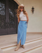 A woman in a sleeveless textured white top, Maebelle Wrap Tie Denim Maxi Skirt, tan cowboy boots, beige cowboy hat, and sunglasses stands on steps before ornate black double doors.