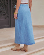 A person wearing the Maebelle Wrap Tie Denim Maxi Skirt, beige cowboy boots, and gold bangles stands on beige stone steps before a dark ornate door, with only their lower body visible.