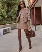A woman stands outdoors wearing a taupe tweed mini dress with a matching jacket, brown knee-high boots, and holding a brown suede handbag.