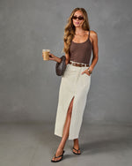 Woman wearing brown ribbed tank top, cream corduroy midi skirt with slit, brown belt, holding iced coffee, black sandals