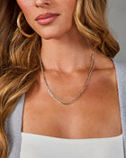 A close-up of a woman wearing a delicate layered necklace with one gold chain and one silver chain, styled with gold hoop earrings and a light gray cardigan.