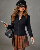 A woman models a black ribbed zip-up sweater with a brown faux-fur collar, paired with a brown pleated skirt, black sunglasses, a black beret, and a black woven handbag against a gray studio background.