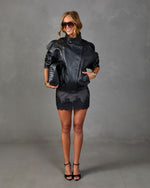 Woman in all-black outfit: leather moto jacket, lace mini skirt, strappy heels, woven clutch, gold bangles, and oversized sunglasses.