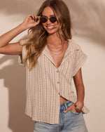 Woman wearing beige striped linen button-up top with jeans, gold jewelry, and oval sunglasses, smiling outdoors