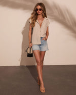 Woman wearing beige striped button-down top, light wash denim cutoff shorts, tan sandals, carrying black bucket bag, sunglasses