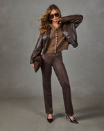 A woman models a stylish brown outfit consisting of a brown faux-leather jacket, a brown button-front knit top, and brown trousers, paired with black heels and sunglasses against a gray studio backdrop.