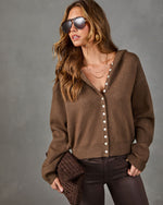 A woman wearing a brown knit button-front cardigan with a hood, styled with dark brown pants, sunglasses, and layered necklaces against a textured gray studio background.
