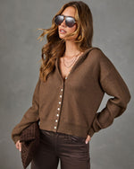 A woman wearing a brown knit cardigan with white buttons, dark brown pants, and sunglasses, holding a brown clutch against a gray studio background.
