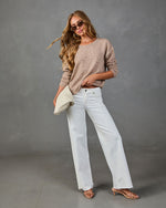 Woman wearing beige knit sweater, white wide-leg jeans, strappy heels, and tortoise sunglasses holding cream clutch bag