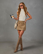 A woman models a beige ribbed knit mock neck top and a matching beige faux-suede mini skirt with front flap pockets. She is styled with knee-high beige suede boots, a cream woven handbag, sunglasses, and holds a coffee cup against a textured gray studio background.