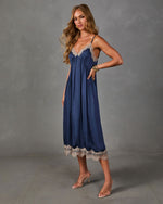Navy satin midi slip dress with beige lace trim, worn with clear-strap heels and gold bracelets against a gray backdrop