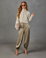 A woman modeling a two-piece outfit consisting of a cream-colored fuzzy mock-neck zip-up sweater and olive green satin pants, styled with sunglasses and clear high-heeled sandals against a gray studio backdrop.