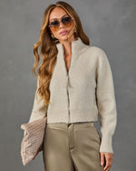 A woman wearing a fuzzy cropped zip-front cardigan in a light oatmeal beige color, paired with khaki pants and holding a textured clutch against a gray studio background.