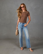 Woman wearing brown short-sleeve knit top, light wash wide-leg jeans, clear heels, holding woven beige clutch bag
